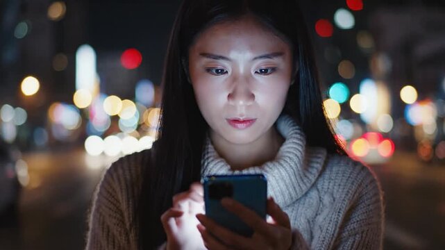 A focused young Asian female worker is using her smartphone at night. The dark city lights create a bokeh background. She interacts with her device late into the evening