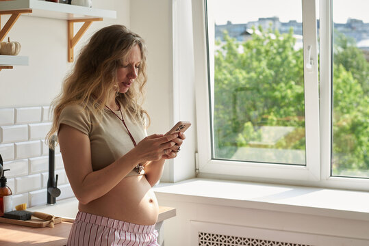 Pregnant woman checks her phone while standing by the window
