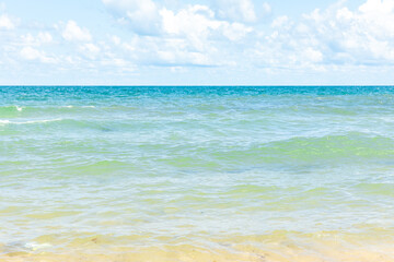 Fototapeta premium Tranquil beach scene with clear turquoise water, gentle waves, and a sandy shoreline under a bright blue sky with fluffy white clouds. Summer vacation and travel concept. Copy space