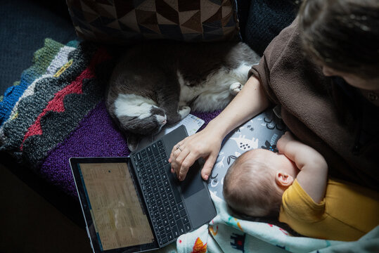 A cat sleeps next to a mother and baby on the couch while she works