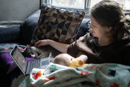 A cat sleeps next to a mother and baby on the couch while she works