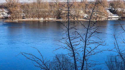 A winter riverbank with snow-covered banks, leafless trees, and an unfrozen river current. A winter river embankment.