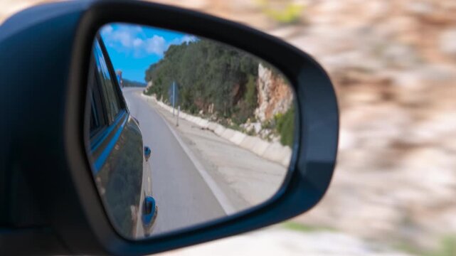 Car side mirror reflecting winding road. Reflection of a beautiful winding mountain road with a bright blue sky in a car's side-view mirror while driving, showing the asphalt and rocky landscape