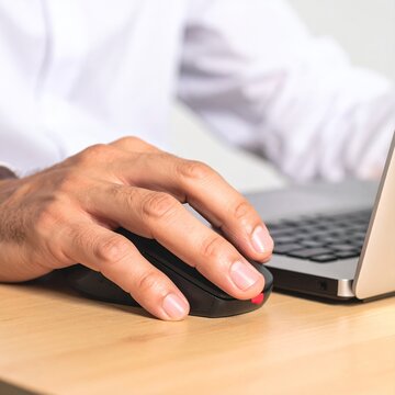 Close-up of a persons hand using a computer mouse next to a laptop on a wooden desk, highlighting digital work and technology interaction.