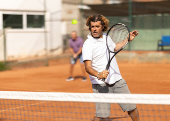 Two players, an adult and an elderly man, play tennis together, they actively move around the court with rackets. Elderly father playing outdoor tennis with his son