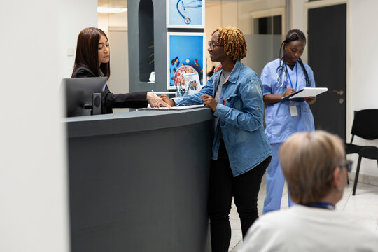 Friendly receptionist at front desk assists patient with updating health records and scheduling treatment visits in clinic. Asian female administrator guiding woman with hospital registration forms.