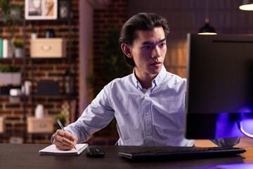 Focused male individual sits at table with computer and notebook, concentrating on freelance work....