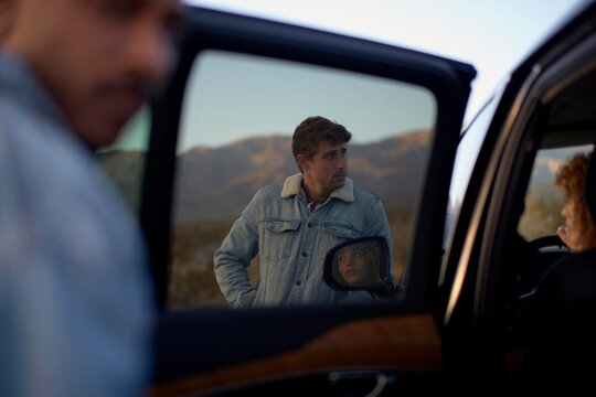Man in Denim Jacket Interacts With Others Next to a Car at Sunset