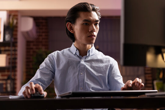 Young male entrepreneur typing and reviewing strategy plans on desktop pc, dedicated on achieving business goals. Asian man works from home at night, completing unfinished tasks and reaching deadlines