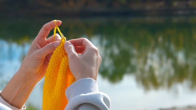 Woman's hands knitting yellow wool outdoors. Close-up of female hands knitting a yellow wool garment using needles outdoors. The tranquil water of a lake or pond provides a soft-focus background