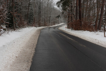 Asfaltowa droga w zimowy dzień. Jezdnia jest czarna, po obu stronach jest wysoki las. Ziemia i gałęzie drzew pokrywa warstwa śniegu. © boguslavus