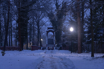 Park w zimowy wieczór. Panuje mrok rozświetlany światłem latarni. Alejki, drzewa, elementy architektury przykrywa warstwa śniegu. © boguslavus