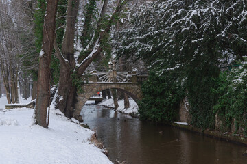 Rzeka Czerna Mała płynąca przez park dworski w mieście Iłowa w Polsce. Nad rzeką widać mosty i kładki dla pieszych. Jest zima, ziemię pokrywa śnieg.