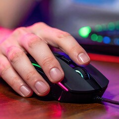 Close up on a left hand gaming with a black computer mouse with RGB lighting on a wooden desk with colorful blurred background gaming setup