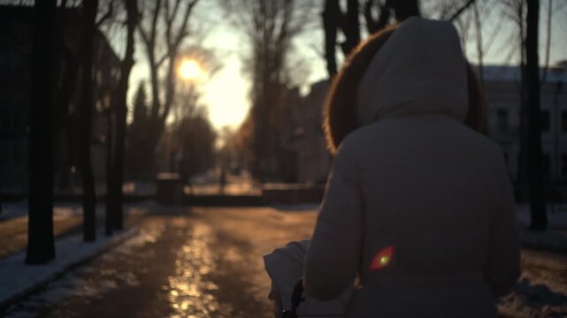 Rear view of a woman wearing a winter coat walking through a park at sunset, pushing a baby stroller