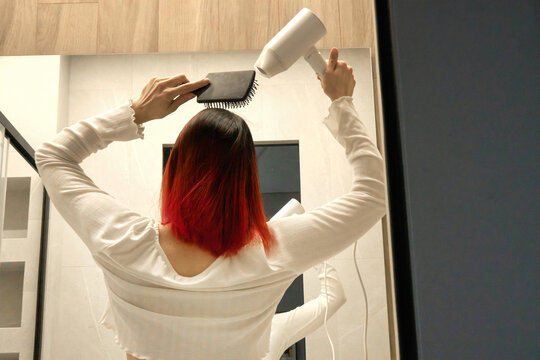 Girl in white shirt with bright red locks drying and styling her hair.