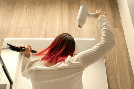 Girl in white shirt with bright red locks drying and styling her hair.