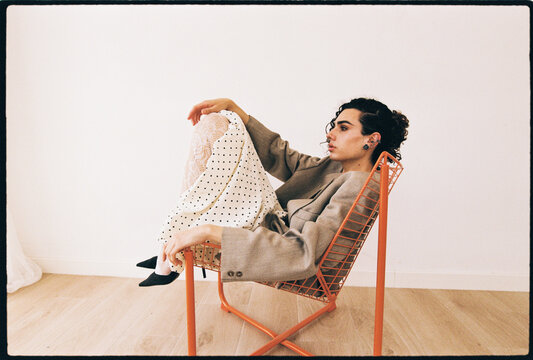 Man posing on orange metal chair wearing blazer and polka dot skirt