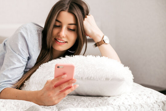 Young smiling woman lying in white bed and using a phone in her bedroom. Happy morning.
