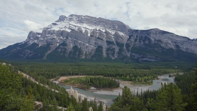 Banff landscape featuring forest, Bow River, hoodoos, and snow capped Mount Rundle under a cloudy sky