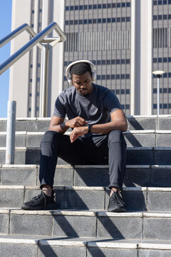 African American man sitting on steps by high-rise, checking watch with headphones and black shoes