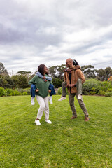 African American family giving piggyback rides on grassy lawn under cloudy sky, wearing sweaters