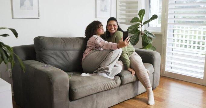 Female friends painting hearts under eyes, snapping selfies, reviewing photos and laughing on couch