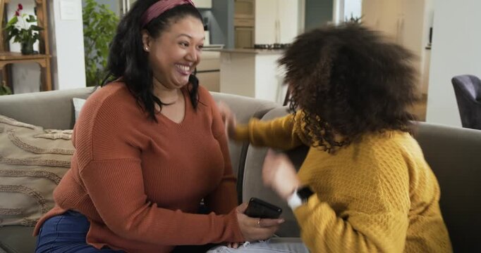 Smartphone tilt sparking mother and daughter laughing, high-fiving, hugging on gray sectional sofa