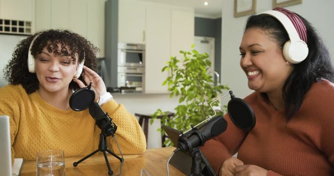 Two women co-hosts adjusting mics, tapping table to jingle and recording discussion in kitchen