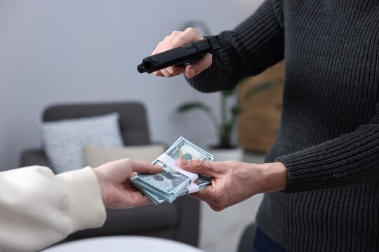 Woman giving money to dangerous criminal with gun indoors, closeup. Armed robbery