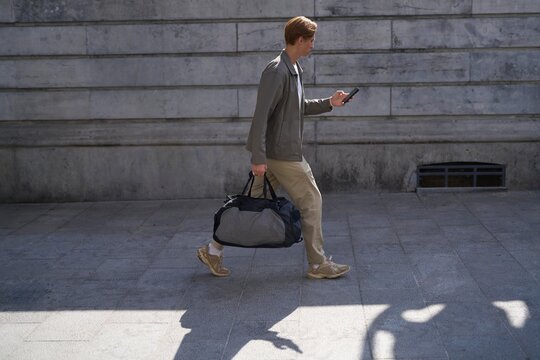 Man Walks on Sidewalk While Checking Phone With Duffel Bag