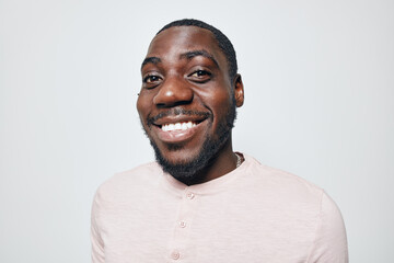 Obraz premium African American man with dark skin smiling and looking at camera in casual light pink shirt. Portrait of happy black male with beard and short hair on white background.