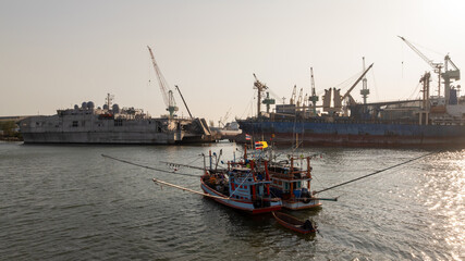 Traditional wooden Thai fishing boat with massive industrial and cargo terminal, Contrast and coexistence between local livelihoods and growth modern heavy industry.