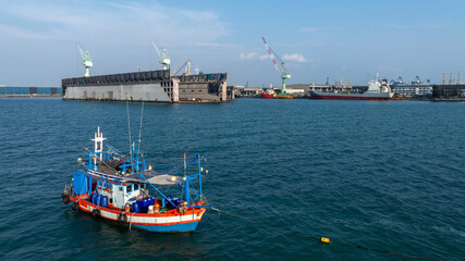 Fototapeta premium Traditional wooden Thai fishing boat with massive industrial and cargo terminal, Contrast and coexistence between local livelihoods and growth modern heavy industry.