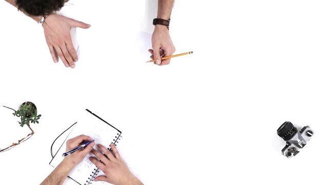 Two hands sketching ideas on a notepad while a camera and a small plant are placed on a bright, minimalist workspace.