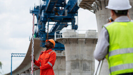 The survey team is operating a theodolite while referencing road construction plans. Civil engineers are taking measurements with surveying equipment at the construction site.