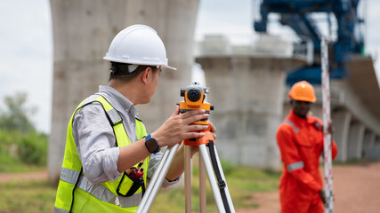 The survey team is operating a theodolite while referencing road construction plans. Civil engineers are taking measurements with surveying equipment at the construction site.