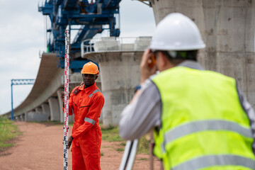 The survey team is operating a theodolite while referencing road construction plans. Civil engineers are taking measurements with surveying equipment at the construction site.