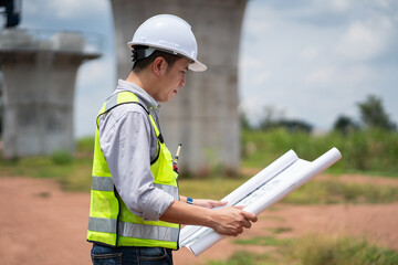 The survey team is operating a theodolite while referencing road construction plans. Civil engineers are taking measurements with surveying equipment at the construction site.