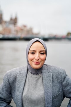 Woman in a Gray Hijab and Blazer Poses by the Water in a City Setting