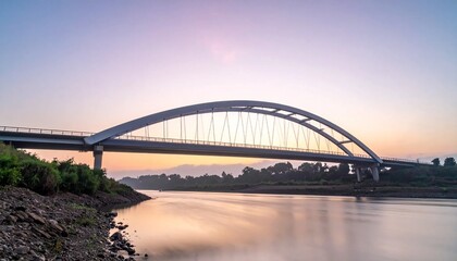 Fototapeta premium Modern Arch Bridge Spanning a Wide River at Sunset with Reflective Waters and Clear Sky.