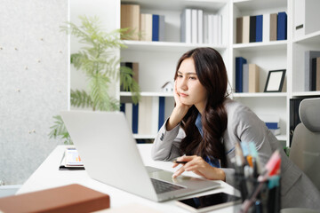 Fototapeta premium A woman is sitting at a desk with a stack of papers in front of her. She is looking at a piece of paper and she is in a state of confusion or frustration 
