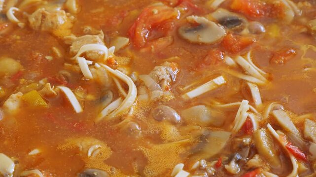 Vegetables with chicken and udon are simmering in a deep cast-iron skillet. Medium shot.