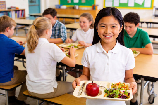 Young smiling Asian student holds tray with apple and salad in school cafeteria. Other children eat in background, showcasing school meal program and nutrition education.