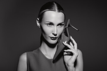 Obraz premium White woman with fair skin and slicked hair holding high heel shoe near face in black and white portrait studio photo.