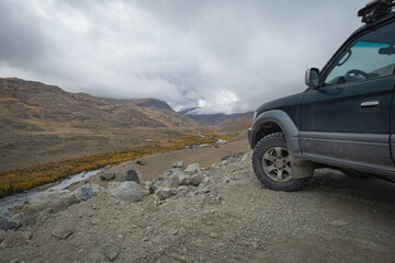 A dark off-road vehicle parked on a dirt mountain road overlooking a winding river valley under a cloudy sky in autumn. © ANDREY