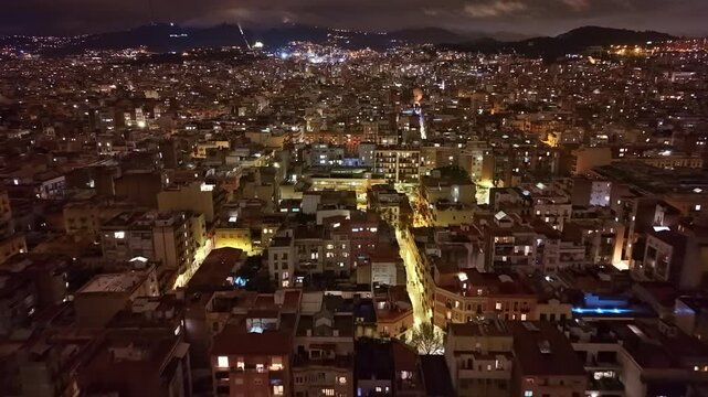 Wide nighttime aerial of Barcelona showing dense urban blocks, glowing arterial streets, and continuous city grid extending toward the Collserola hills, capturing the energy of the Mediterranean city