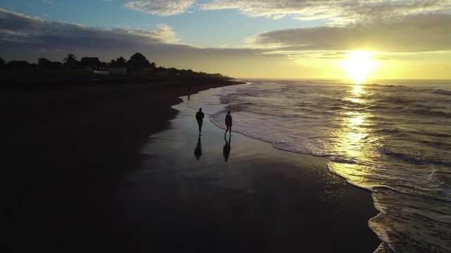 Aerial zoom in of a couple walking by the shoreline during sunset at Los Cristianos beach in Tenerife with reflections on wet sand.