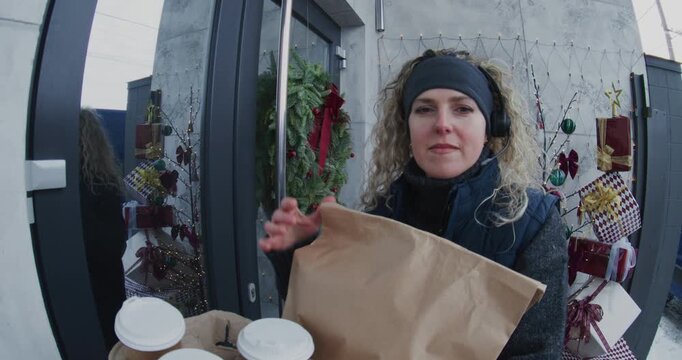 Female Courier With Headset Delivers Hot Coffee Cups and Brown Paper Bag to Man Doorstep, Interaction Captured by Smart Doorbell Camera near Winter Decorated House. Concept of Modern Service Delivery.