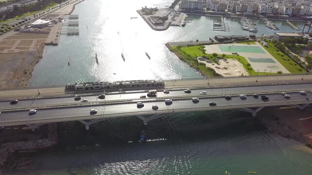 Aerial drone shot capturing two tramway trains meeting on the Hassan II bridge. Features the modern Marina Sal&eacute;, river traffic, and busy vehicle lanes in a sunny Mediterranean landscape.